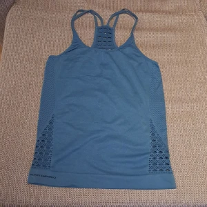 Blå träningstopp från Armani - Säljer en snygg träningstopp. Den är ärmlös och har en tight passform, perfekt för gymmet eller yoga. Toppen har ett coolt mönster med meshdetaljer på sidorna och ryggen, vilket ger bra ventilation. Materialet är mjukt och stretchigt, per. f. träning
