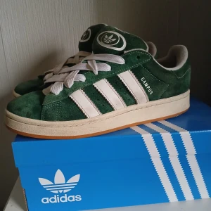 Gröna Adidas Campus sneakers - Säljer ett par snygga gröna Adidas Campus sneakers i mocka. De har de klassiska vita ränderna på sidan och en vit sula. Skorna är i bra skick och kommer med extra gröna skosnören. Perfekta för både vardag och fest!