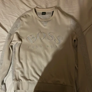Beige tröja från Hugo Boss - Säljer en snygg beige tröja från Hugo Boss. Den har ett stort BOSS-tryck på framsidan och är superbekväm. Perfekt för både vardag och lite finare tillfällen. Tröjan har långa ärmar och en klassisk rund halsringning. Passar perfekt till jeans eller chinos!