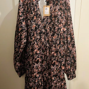 Blommig klänning från Shein - Säljer en superfin blommig klänning från Shein i storlek XL. Den har en svart bas med ett vackert blommönster i rosa och vita nyanser. Klänningen är långärmad och perfekt för både höst och vår. Materialet känns lätt och bekvämt, vilket gör den idealisk för både vardag och festligare tillfällen.