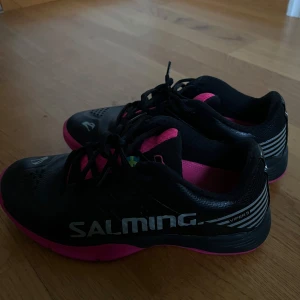 Svarta och rosa Salming sneakers - Säljer ett par snygga Salming sneakers i svart och rosa. Skorna har en sportig design med snörning och Salming-loggan på sidan. De är perfekta för träning eller vardagsbruk. Skorna är i bra skick och redo för nya äventyr! Köpt för 899 säljer 699