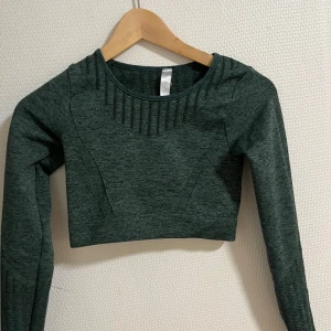 Mörkgrön croppad träningstopp - Säljer en snygg mörkgrön croppad träningstopp med långa ärmar. Toppen har en ribbad detalj vid halsen och är perfekt för gymmet eller yoga. Materialet är stretchigt och bekvämt, vilket gör den idealisk för träning. Passar perfekt till ett par höga tights!