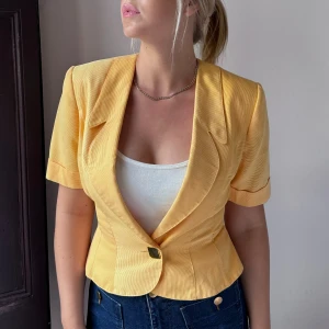 Gul kavaj med kortärmad design - Säljer en superfin gul kavaj med korta ärmar. Den har en snygg knapp framtill och en figursydd passform som ger en elegant look. Köpt vintage i Barcelona. Står stl. 42 i kavajen, men skulle säga att den är 38