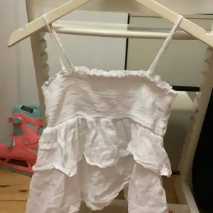 Vit volangtopp från Gina Tricot - Söt vit volangtopp från Gina Tricot med smala axelband som är regleringsbara och volanger. Perfekt för varma sommardagar. Toppen har en elastisk överdel för en bekväm passform.