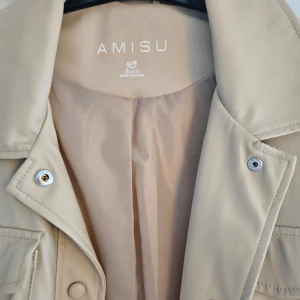 Beige skinn jacka från Amisu - Snygg beige skinn jacka från Amisu med knappar och bälte i midjan. Jackan har långa ärmar och flera fickor framtill, vilket ger en stilren look. Perfekt för vår och höst!