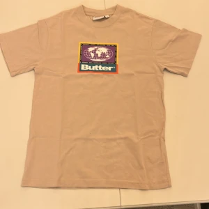 Beige t-shirt från Butter - Säljer en beige t-shirt från Butter med ett färgglatt tryck på framsidan. Skriv om ni har frågor.