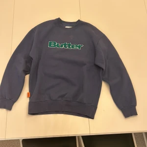Blå tjocktröja från Butter - Säljer en blå sweatshirt från Butter med grönt och vitt tryck på bröstet. Tröjan har långa ärmar. Skriv om ni har frågor.