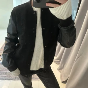 Svart bomberjacka med skinnärmar - Snygg svart bomberjacka med skinnärmar och knappar framtill. Perfekt för en cool och avslappnad stil. Jackan har en klassisk design med ribbade muddar vid ärmslut.