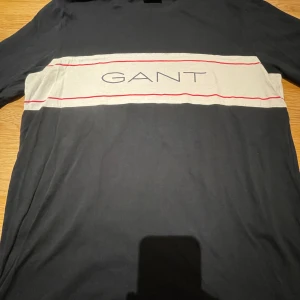 Svart långärmad tröja från GANT - Snygg svart långärmad tröja från GANT med ett brett vitt band och röda linjer över bröstet. GANT-loggan är tryckt i mitten av det vita bandet. Perfekt för en stilren och avslappnad look.
