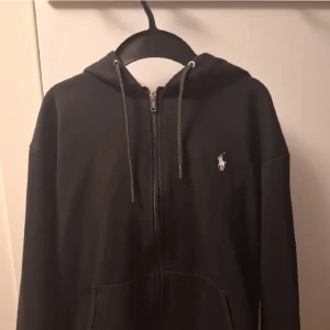 Svart hoodie från Ralph Lauren - Säljer en stilren svart hoodie från Ralph Lauren med dragkedja och det klassiska logotypbroderiet på bröstet. Perfekt för en avslappnad look.
