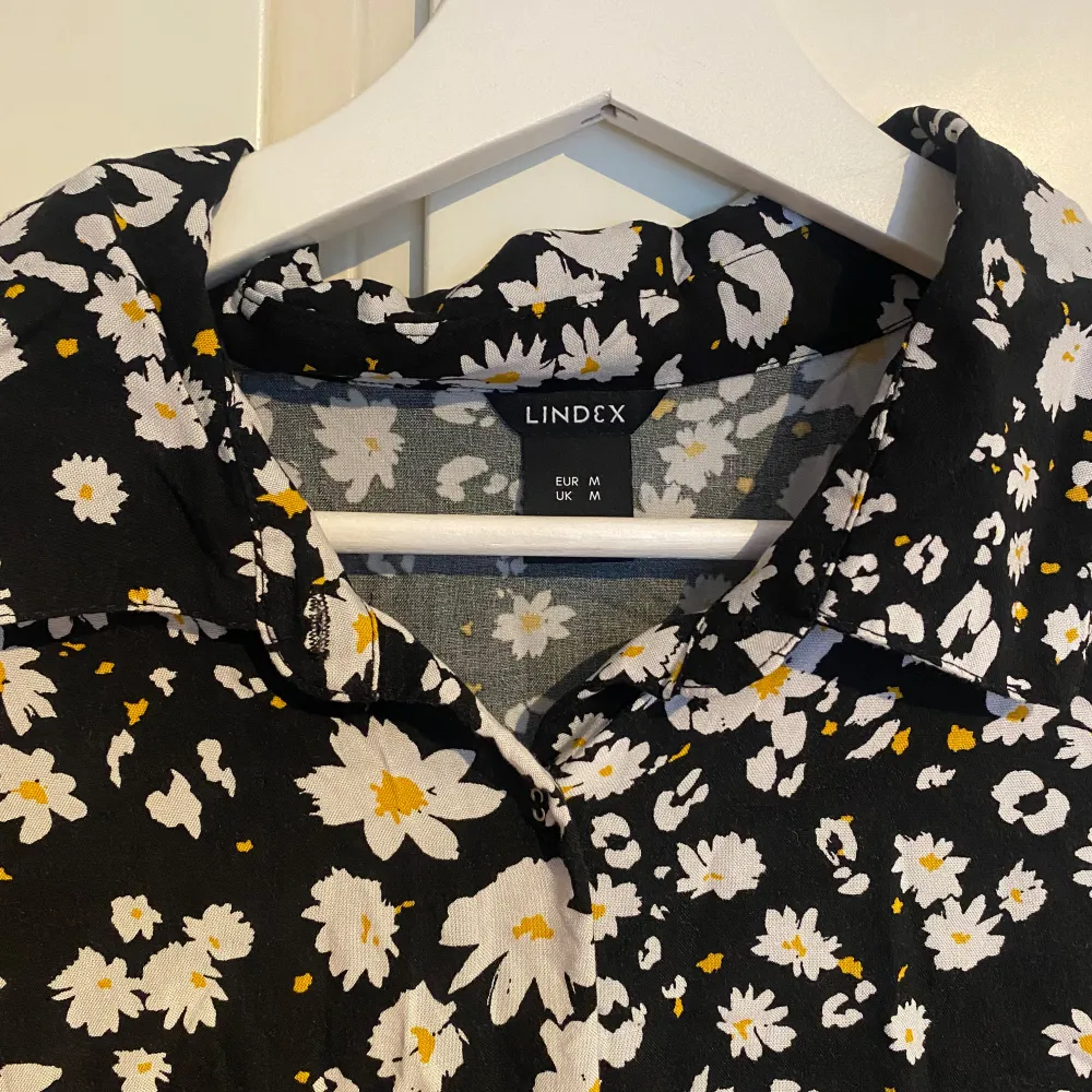 Svart klänning från Lindex med ett blommigt mönster i vitt och gult. Klänningen har långa ärmar och ett knytband i midjan samt knappar i fram.. Mekot.