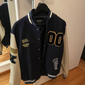 Blå och vit bomberjacka från MAN - Snygg blå och vit bomberjacka från boohoo med tryck och broderade detaljer. Jackan har beigea ärmar med blåa detaljer och en ribbad krage. Perfekt för en cool och avslappnad stil.