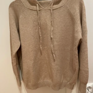 Merinoull hoodie - Säljer en stilren beige merinoull hoodie med långa ärmar och justerbar huva. Perfekt för en avslappnad look. Ganska kort i storleken