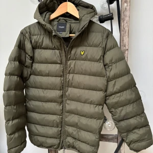 Olivgrön dunjacka från Lyle & Scott - Snygg olivgrön pufferjacka från Lyle & Scott junior med huva och dragkedja. Den ikoniska loggan på bröstet. Jackan har en bekväm passform, passar både kille & tjej.