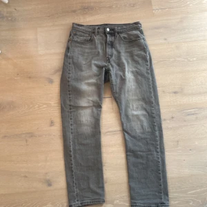 Grå jeansbyxor från Levi's - Säljer ett par grå jeansbyxor från Levi's med klassisk femficksdesign och knappgylf. Byxorna har en rak passform och är perfekta för en avslappnad stil. Levi's logotyp syns på bakfickan. 