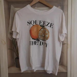 Vit t-shirt med tryck från FB Sister - Säljer en vit t-shirt från FB Sister med ett färgglatt tryck av en apelsin och texten 'Squeeze the Day'. T-shirten har korta ärmar och en rund halsringning. (den är i storlek xl men känns mindre, mer som en M) 