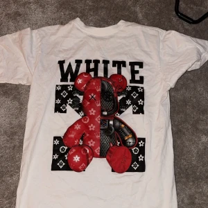 Vit t-shirt med tryck från Off-White - Snygg vit t-shirt från Off-White med ett unikt tryck av en röd och svart nalle på baksidan och texten 'WHITE'. Framsidan har en röd text 'OFF'. Perfekt för en trendig look.