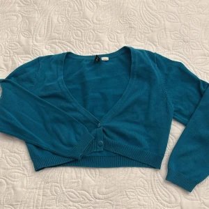 Blå kofta från H&M Divided - Säljer en snygg blå kofta från H&M Divided. Den är croppad med en v-ringad design och knappar framtill.