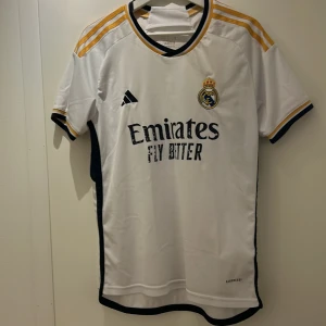 Vit Real Madrid fotbollströja från Adidas - Säljer en vit Real Madrid fotbollströja från Adidas med korta ärmar och gula detaljer. Tröjan har klubbens emblem broderat på bröstet och sponsorlogga framtill. Perfekt för alla fans av laget! ⚽
