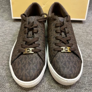 Bruna sneakers från Michael Kors - Snygga bruna sneakers från Michael Kors med ett diskret mönster och gyllene detaljer. Skorna har en vit sula och klassisk snörning. Perfekta för en stilren look i storlek 37,5. Använda endast 2 gånger. 