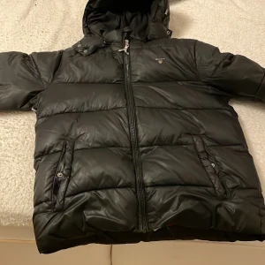 Svart dunjacka från GANT - Snygg svart dunjacka från GANT med huva och dragkedja framtill. Jackan har en röd insida och praktiska fickor med knappar. Perfekt för kalla dagar. Jackan är för nån som är 170 funkar på andra också själv är 178 och den passar fortfarande 