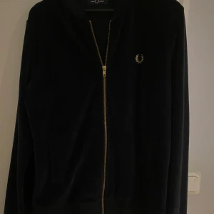 Svart jacka från Fred Perry - Snygg svart jacka från Fred Perry med dragkedja och guldfärgad logga på bröstet. Jackan har långa ärmar och en stilren design, perfekt för en casual look.