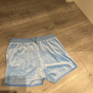 Ljusblå velourshorts från Juicy Couture - Säljer ett par ljusblå velourshorts från Juicy Couture med snörning i midjan och en broderad logga. Shortsen har en bakficka och är supermjuka och bekväma.