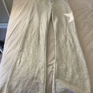 Grå mjukisbyxor från Kappahl - Säljer ett par grå mjukisbyxor från Kappahl med en vit stjärndetalj på benet. Byxorna har en bekväm passform och är perfekta för en avslappnad stil. De är tillverkade i ett mjukt material som känns skönt mot huden.