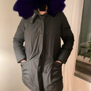 Svart jacka från Woolrich - Säljer en stilren svart jacka från Woolrich. Jackan har långa ärmar och knappar framtill. Den är perfekt för kyligare väder och har en klassisk design med praktiska fickor. OBS! Givetvis med den lurviga luvan!