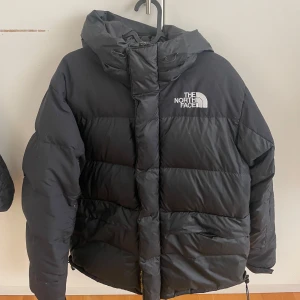 Svart dunjacka från The North Face - Säljer en svart dunjacka från The North Face med huva och dragkedja. Jackan är perfekt för kalla dagar och har en klassisk pufferdesign med logotyp på bröstet och på ryggen.