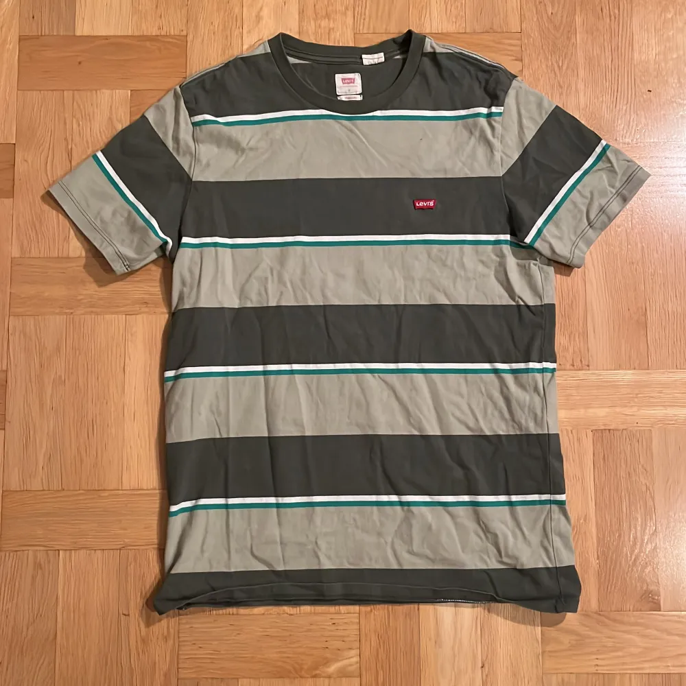 Snygg randig t-shirt från Levi's i gröna och gråa nyanser med vita och turkosa detaljer. Perfekt för en avslappnad stil. Den har korta ärmar och en klassisk passform.. T-paidat.