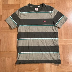 Randig t-shirt från Levi's - Snygg randig t-shirt från Levi's i gröna och gråa nyanser med vita och turkosa detaljer. Perfekt för en avslappnad stil. Den har korta ärmar och en klassisk passform.