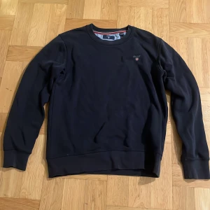 Svart sweatshirt från Gant - Säljer en stilren svart sweatshirt från Gant med rund hals och diskret logga på bröstet. Tröjan har långa ärmar och ribbade muddar vid ärmslut och nederkant. Perfekt för en avslappnad look.