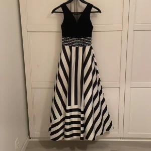 Svart och vit långklänning från Pery Bella - Klänningen ser helt nytt utan, det är bara 2 vita prickar på framsidan som saknas. Elegant svart och vit långklänning från Pery Bella med ett randigt mönster och glittriga detaljer i midjan. Midjan är dekorerad med ett stilrent mönster av vita prickar. Klänningen har en v-ringad topp och är ärmlös, vilket ger en stilren look. Perfekt för speciella tillfällen. 