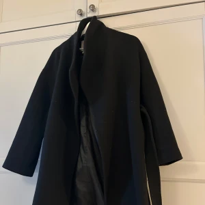 Svart omlottkappa  - Elegant svart kappa från hm med en stilren design. Omlottvariany med skärp i midjan och lite större slag. Knälång. Inga större anmärkningar, i gott skick.