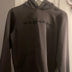 Grå hoodie från Sail Racing - Snygg grå hoodie från Sail Racing med svart logotyp på bröstet. Tröjan har en klassisk design med huva och långa ärmar. Perfekt för en avslappnad stil.