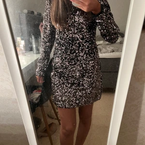 Svart glittrig klänning med öppen rygg - Snygg svart klänning täckt av glittriga paljetter. Den har en öppen rygg med en dekorativ rosett och långa ärmar. Perfekt för en festlig kväll! Prislapp kvar och den är i storlek 158/164 men passar mig med Xs