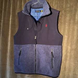 Fleece Väst - Ralph Lauren - Säljer en snygg blå fleeceväst från Polo Ralph Lauren med dragkedja och två fickor framtill. Västen har en broderad röd logotyp på bröstet och en hög krage för extra värme. Perfekt för kyliga dagar! Vid minsta fråga eller fundering hör av er🤝