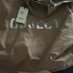Beige shoppingväska från Object - Snygg och rymlig beige shoppingväska från Object med texten 'MADE BY PEOPLE FOR PEOPLE'. Perfekt för att bära med sig allt man behöver under dagen. Väskan har en praktisk ficka på utsidan.