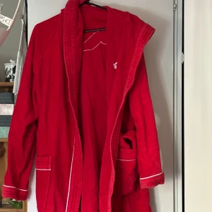 Röd morgonrock från Puma - Säljer en röd morgonrock från Puma med vit piping och logga på ryggen. Den har långa ärmar och praktiska fickor framtill. Perfekt för att mysa hemma eller efter träningen.