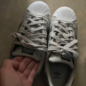 Adidas Superstar sneakers - Snygga Adidas Superstar sneakers i vitt med de klassiska tre ränderna i ett unikt nätmönster. Skorna har snörning och den ikoniska tåhättan i gummi. Perfekta för en stilren look.