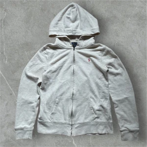 Grå Ralph Lauren Hoodie  - Säljer en stilren grå hoodie från Polo Ralph Lauren med dragkedja och huva. Den har en liten rosa logga på bröstet och är tillverkad i mjukt material. Perfekt för en avslappnad look.