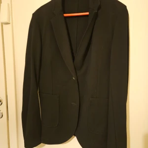 Svart kavaj från J.Lindeberg - Elegant svart kavaj från J.Lindeberg i en blandning av viskos och polyamid. Kavajen har en klassisk design med knappar framtill och fickor på sidorna. Perfekt för en stilren look.