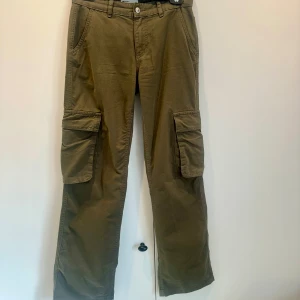 Olivgröna cargobyxor - Säljer ett par olivgröna cargobyxor med flera praktiska fickor från Gina tricot i strl 32. Byxorna har en normal passform. De är tillverkade i ett slitstarkt material och har en klassisk cargodesign med fickor på sidorna och bak.