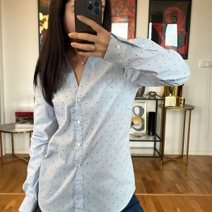 Ljusblå prickig skjorta - Säljer en superfin ljusblå skjorta med små prickar. Den har en klassisk krage och knappar hela vägen ner. Skjortan är långärmad och perfekt för både vardag och fest. Materialet känns som bomull och den har en normal passform. Perfekt för vår och sommar!