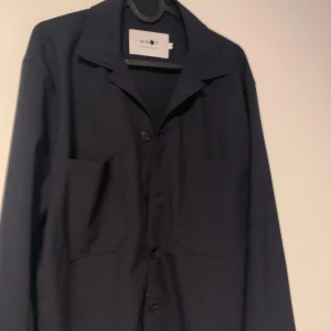 Svart overshirt från NN07 - Säljer en stilren marinblå overshirt från NN07 i storlek S. Den har en klassisk krage och knäppning framtill med stora fickor. Perfekt för lager-på-lager under hösten eller våren. Passar både till vardags och lite mer uppklädda tillfällen.