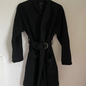 Svart kappa från Forever 21 - Säljer en stilren svart kappa från Forever 21. Den har ett bälte i midjan för en snygg passform och stora fickor framtill. Perfekt för höst och vår!