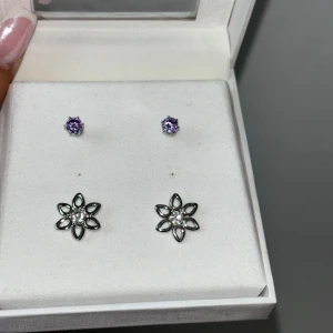 Snygga örhängen i lila och silver - Säljer två par örhängen köpta på Punktshop. Ger en extra touch till din outfit, både till vardags och fest. De lila stenarna är små och gnistrande, medan de blommiga örhängena har en elegant design med små pärlor i mitten.