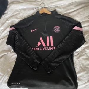 Svart långärmad fotbollströja från Nike - Säljer en svart långärmad fotbollströja från Nike med rosa detaljer. Tröjan har en halv dragkedja framtill och är tillverkad i Dri-FIT material för att hålla dig torr och bekväm. Perfekt för träning eller casual wear. PSG-logga på bröstet ger en extra touch av stil.