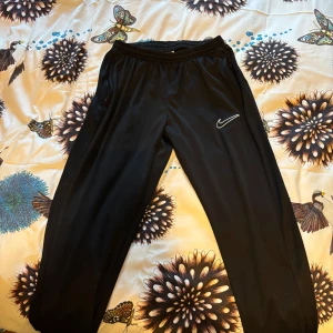 Svarta träningsbyxor från Nike - Säljer ett par svarta träningsbyxor från Nike i storlek L. Byxorna är tillverkade i ett lätt och bekvämt Dri-FIT material som håller dig torr under träningen. De har en elastisk midja och den klassiska Nike-loggan broderad på sidan. Perfekta för gymmet eller löprundan!